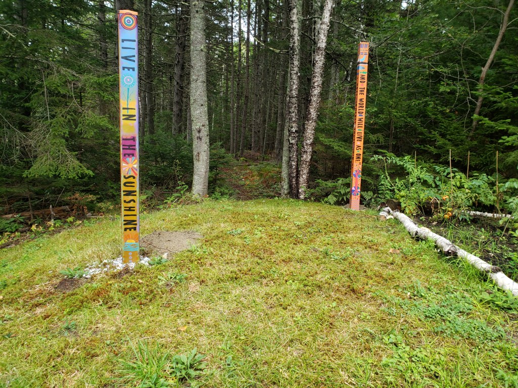 Grassy knoll punctuated by two colorful Poetry Poles framing the entrance into our spruce wood where there is the remains of a an old granite quarry now covered in moss.
